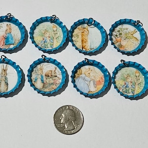 Könnte beinhalten: Acht blaue Flaschenverschluss-Anhänger mit Illustrationen von Peter Hase und anderen Figuren. Jeder Verschluss hat eine kleine Schlaufe zum Aufhängen. Eine Münze dient als Größenreferenz.