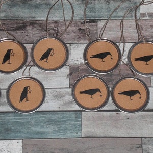 May include: A collection of ten round ornaments with a metal rim and a brown center. Each ornament features a black crow silhouette holding a star, suspended by twine. The background is a rustic wooden surface.