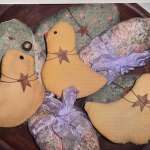 May include: A wooden bowl holds three yellow fabric bird ornaments with star accents and red beaks. The birds are surrounded by fabric-wrapped shapes with floral patterns and tied with lavender ribbons. A rustic, country-style decorative item.