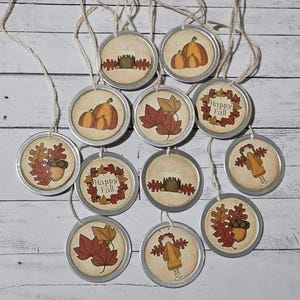 May include: A collection of round, rustic-style ornaments with various fall-themed designs. Each ornament features a metal rim and a printed image of pumpkins, leaves, or a candle. Some ornaments have the text "Happy Fall". They are strung with twine.