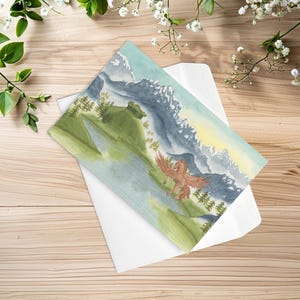 May include: A rectangular greeting card featuring a watercolor illustration of a brown winged horse standing in a green landscape with a lake, pine trees, and snow-capped mountains under a pale sky, resting on a white envelope.