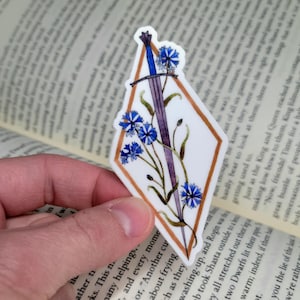 Op de afbeelding: Een sticker met een blauw zwaard en blauwe bloemen in een witte ruit met een gouden rand. De sticker wordt in een hand gehouden, met een boek op de achtergrond. Fantasiethema.