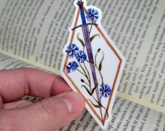 May include: A sticker featuring a blue sword with a blue floral design. The sword is set against a white diamond shape with a gold border. The sticker is held in a person's hand, with a book in the background.