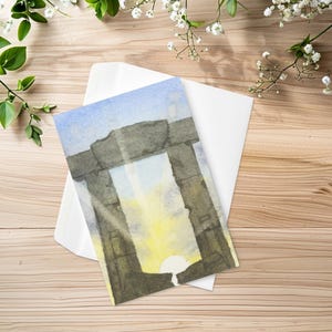 May include: A rectangular greeting card featuring a watercolor illustration of a stone trilithon structure. The artwork depicts a bright yellow sun rising between the dark gray stone pillars against a light blue sky, with a white envelope underneath.