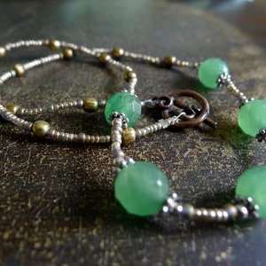 May include: A delicate necklace featuring faceted green beads interspersed with smaller metallic beads. The necklace has a decorative clasp and is displayed on a textured, brown surface. The beads are approximately 1.3 cm in diameter.