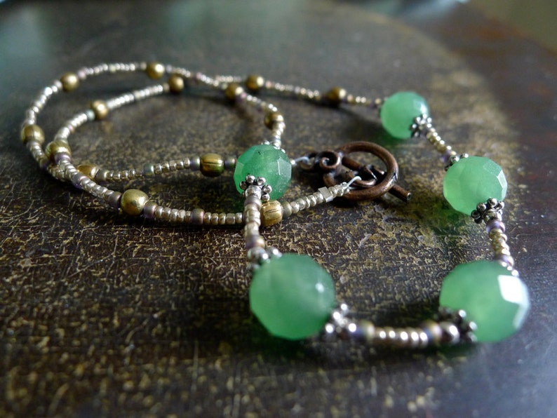 May include: A delicate necklace featuring faceted green beads interspersed with smaller metallic beads. The necklace has a decorative clasp and is displayed on a textured, brown surface. The beads are approximately 1.3 cm in diameter.