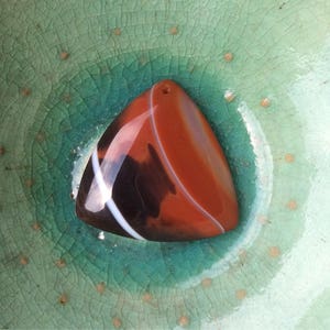 May include: A triangular, polished gemstone pendant with a hole for stringing. The stone features bands of dark brown, white, and reddish-orange hues. The pendant is displayed on a textured, teal-colored surface.