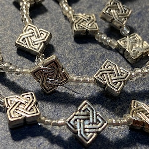 Könnte beinhalten: Nahaufnahme einer Halskette mit quadratischen, silberfarbenen Perlen mit keltischen Knotenmustern. Die Perlen sind mit kleinen, klaren Glasperlen durchsetzt, die auf einer zarten Schnur aufgereiht sind. Der Hintergrund ist ein gedämpftes Blau.