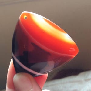 Op de afbeelding: Een gepolijste, driehoekige hanger met een gat aan de bovenkant. De steen heeft banden van rood, oranje, zwart en wit. De hanger wordt in de hand van een persoon gehouden. De steen is glad en lijkt een soort agaat te zijn.