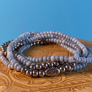Op de afbeelding: Een stapel armbanden met grijze en zilverkleurige kralen. De armbanden zijn gemaakt van kleine, ronde kralen. Eén armband heeft een zilveren bedel met de tekst "HOPE". De armbanden liggen op een bruin oppervlak met een blauwe achtergrond.