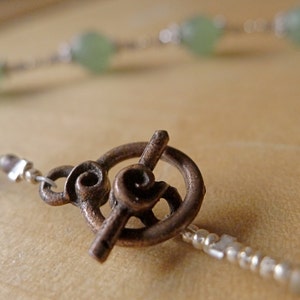 May include: Close-up of a necklace with a bronze toggle clasp. The necklace features small silver beads and light green, round beads. The clasp has a decorative spiral design. The background is a blurred, neutral tone.