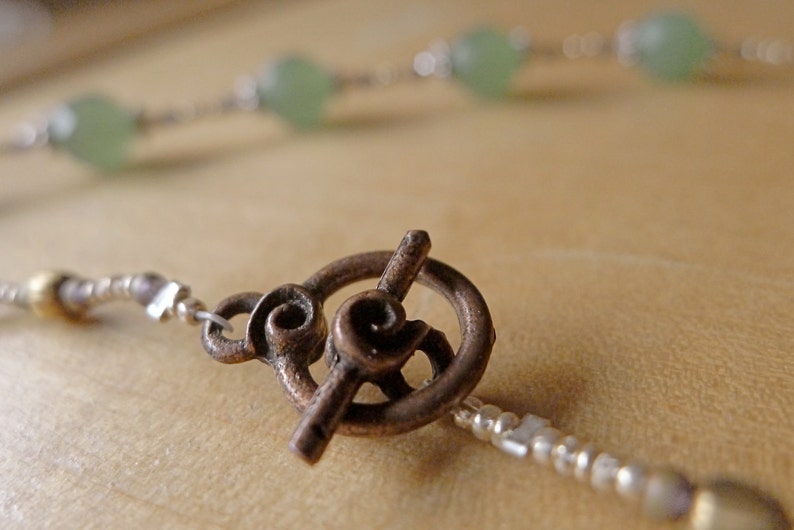 May include: Close-up of a necklace with a bronze toggle clasp. The necklace features small silver beads and light green, round beads. The clasp has a decorative spiral design. The background is a blurred, neutral tone.
