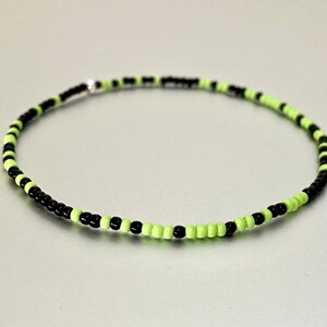 May include: A beaded anklet featuring alternating black and neon green beads. The delicate jewelry piece is strung on a thin, flexible cord and has a small silver-colored clasp. The anklet is designed to be worn around the ankle.