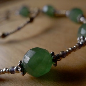 May include: A close-up of a necklace featuring large, faceted, green gemstone beads. The beads are interspersed with smaller silver and gold-toned beads. The necklace is displayed on a wooden surface.