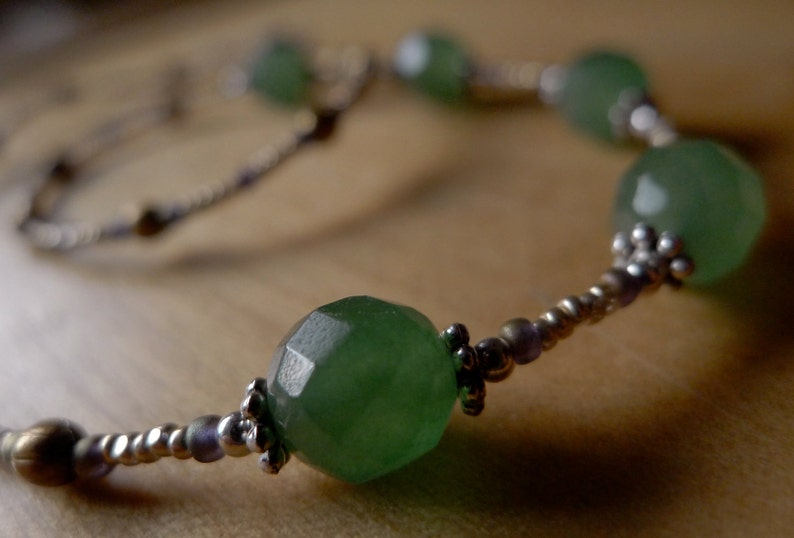 May include: A close-up of a necklace featuring large, faceted, green gemstone beads. The beads are interspersed with smaller silver and gold-toned beads. The necklace is displayed on a wooden surface.