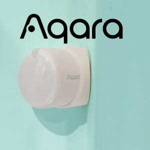 May include: A white Aqara smart home device fixed to a light blue wall. The device features a rounded front and a rectangular base. The Aqara logo is displayed in black at the top and in grey on the device.