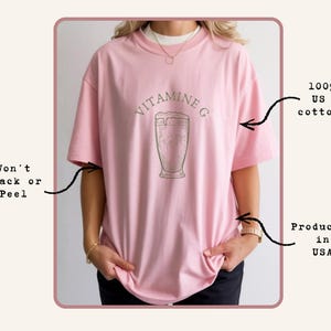 May include: A light pink t-shirt with the text "VITAMINE G" above a graphic of a beverage in a glass. The shirt is made of 100% US cotton and is produced in the USA.