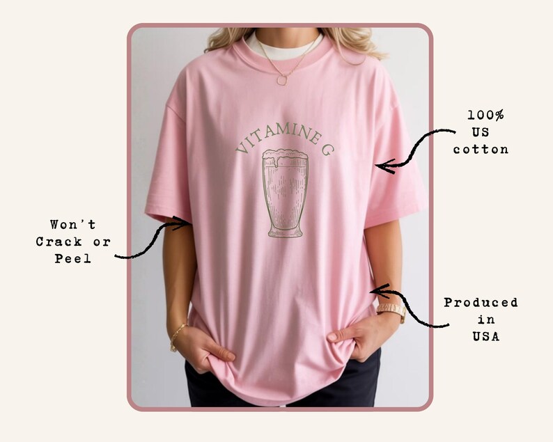 May include: A light pink t-shirt with the text "VITAMINE G" above a graphic of a beverage in a glass. The shirt is made of 100% US cotton and is produced in the USA.