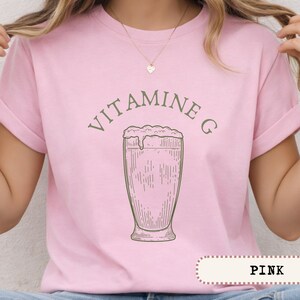 May include: Pink t-shirt with rolled-up sleeves featuring the text "VITAMINE G" above a sketched image of a drink in a tall glass. The word "PINK" is displayed in a banner at the bottom of the shirt.