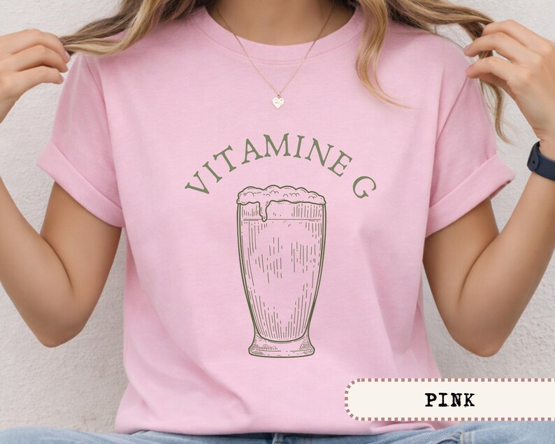 May include: Pink t-shirt with rolled-up sleeves featuring the text "VITAMINE G" above a sketched image of a drink in a tall glass. The word "PINK" is displayed in a banner at the bottom of the shirt.