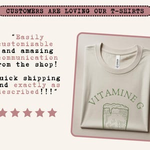 May include: A folded, light beige t-shirt with a crew neck. The shirt features the text "VITAMINE G" in green, along with a green illustration of a glass of beer. The image also includes customer testimonials and the phrase "CUSTOMERS ARE LOVING OUR T-SHIRTS".