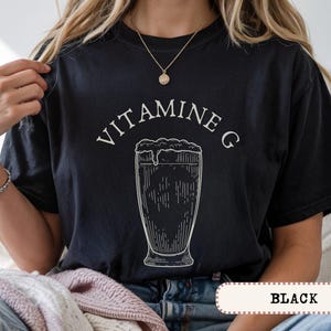 May include: Black t-shirt with the text "VITAMINE G" above a detailed illustration of a beer glass. The shirt features a vintage-style design, with the word "BLACK" at the bottom.