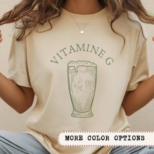 May include: Cream-colored t-shirt with the words "VITAMINE G" arched above a green illustration of a frothy drink in a tall glass. The shirt has short sleeves and a round neckline. The text "MORE COLOR OPTIONS" is at the bottom.