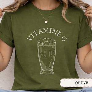 May include: Olive green t-shirt with the text "VITAMINE G" above a detailed line drawing of a beer glass. The shirt is a solid color with rolled-up sleeves.