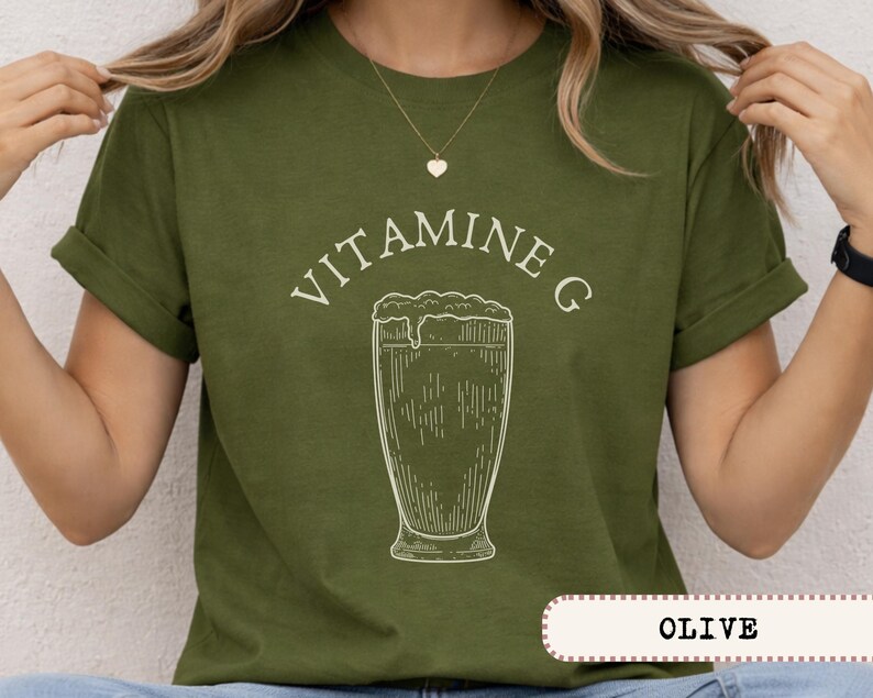 May include: Olive green t-shirt with the text "VITAMINE G" above a detailed line drawing of a beer glass. The shirt is a solid color with rolled-up sleeves.