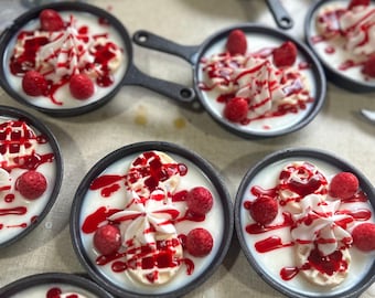 Könnte beinhalten: Eine Sammlung kleiner schwarzer gusseiserner Pfannen, gefüllt mit weißem Wachs, verziert mit Miniaturwaffeln, weißen Schlagsahnehäubchen und roten Beeren, alles übergossen mit einem glänzenden roten Sirup, der an Fruchtsauce erinnert.