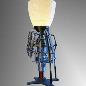 May include: A silver and black rocket engine lamp with a white, textured lampshade. The lamp has a copper base and a black stand with three legs.