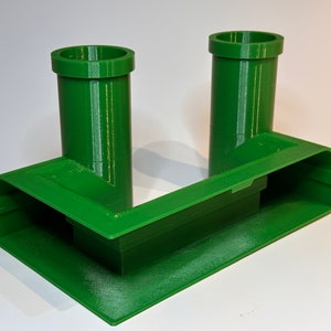 May include: A green 3D printed stand with two vertical pipes, resembling a video game character's iconic pipes.