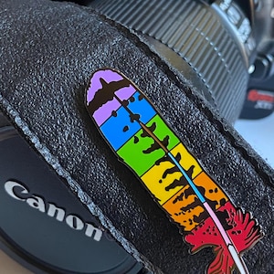 May include: A colorful feather-shaped enamel pin with a rainbow gradient design. The pin is set against a black camera strap and a Canon lens cap. The feather features a spectrum of colors, including purple, blue, green, yellow, orange, and red.