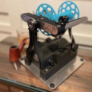 May include: A black and silver yarn winder with a light blue spool. The winder sits on a silver base and has a decorative design on the side. A small, red pipe and a glass of liquid are in the background.