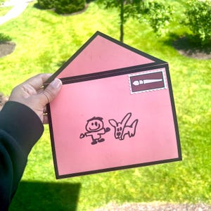 May include: A pink envelope with a black border, featuring a cartoon drawing of a person and a rabbit. A stamp with a paintbrush design is in the upper right corner. The envelope is held against a background of green grass and trees.