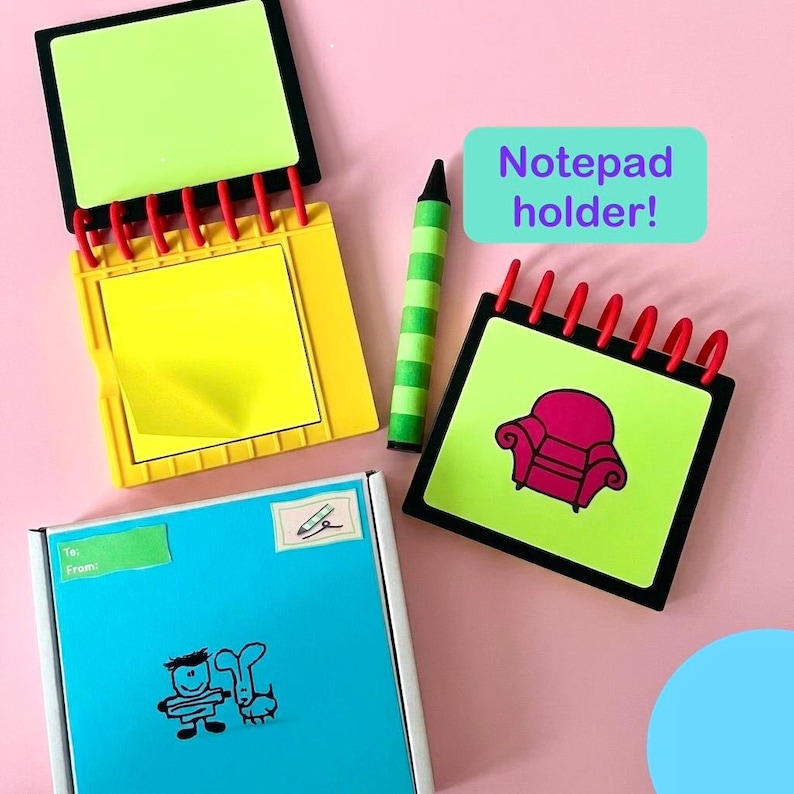 May include: A collection of notepad holders in various designs. One notepad holder is yellow with a black border, another is black with a red sofa graphic, and a third is blue with a cartoon drawing. A green and yellow crayon is also included.