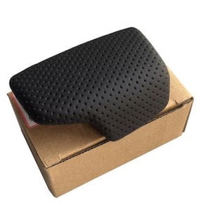 May include: A black, perforated leather automotive part rests on a brown cardboard box. The part has a curved shape and is covered in small, evenly spaced holes. The box is plain, with a simple design.