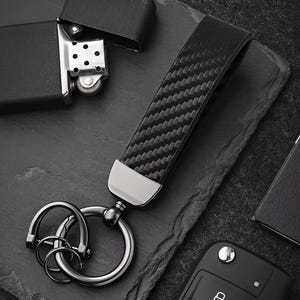 May include: A black carbon fiber key fob with a silver metal ring and keychains. The key fob has a textured pattern and a silver metal accent at the base. A black lighter is in the background.