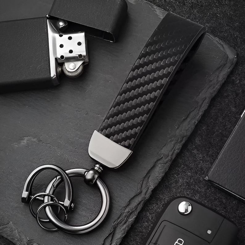 May include: A black carbon fiber key fob with a silver metal ring and keychains. The key fob has a textured pattern and a silver metal accent at the base. A black lighter is in the background.