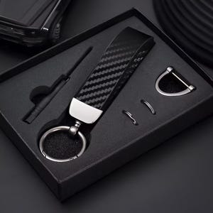 May include: A black carbon fiber keychain set in a black gift box. The keychain has a silver metal accent and a large key ring. The set includes a small tool and two additional metal rings.