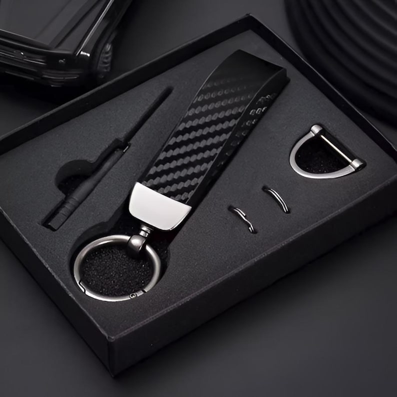 May include: A black carbon fiber keychain set in a black gift box. The keychain has a silver metal accent and a large key ring. The set includes a small tool and two additional metal rings.