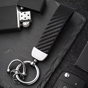 May include: Black leather key fob with a carbon fiber weave pattern. The fob has a silver-colored metal accent and a split key ring. A black lighter and car key fob are also in the image.