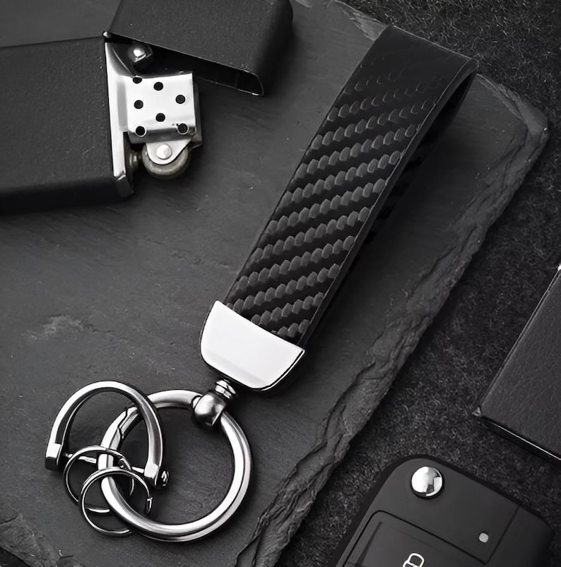May include: Black leather key fob with a carbon fiber weave pattern. The fob has a silver-colored metal accent and a split key ring. A black lighter and car key fob are also in the image.