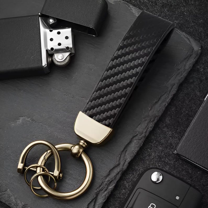 May include: Black carbon fiber key fob with gold-colored hardware. The key fob has a textured pattern and a gold-colored metal ring for keys. A black lighter and car key are also in the image.