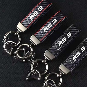 May include: Four keychains with carbon fiber straps and silver-tone hardware. Each strap has the text "RS 3" in white. The edges of the straps are stitched with red thread. The keychains are on a dark background.