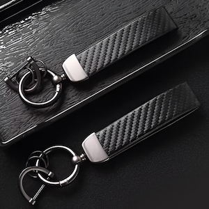 May include: Two black carbon fiber keychains with silver-tone metal accents. The keychains have a woven pattern and a metal clasp. The keychains are on a dark surface.