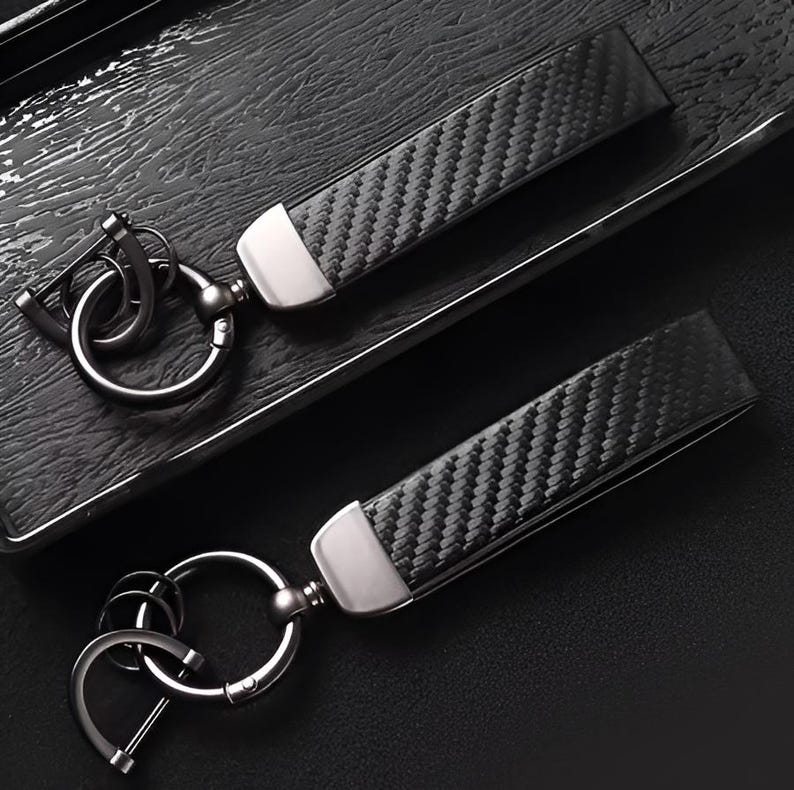 May include: Two black carbon fiber keychains with silver-tone metal accents. The keychains have a woven pattern and a metal clasp. The keychains are on a dark surface.