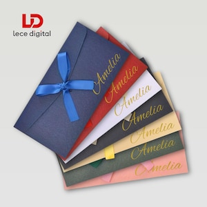 May include: A stack of seven rectangular envelopes in navy, red, white, black, cream, dark green, and light pink. Each envelope features the name Amelia printed in gold script. The top navy envelope is tied with a blue satin ribbon bow.