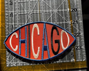 May include: Embroidered patch with the word "CHICAGO" in white, blue, and black lettering on a red oval. The patch is outlined in blue stitching. The patch is placed on a black and gray cutting mat with measurement markings.