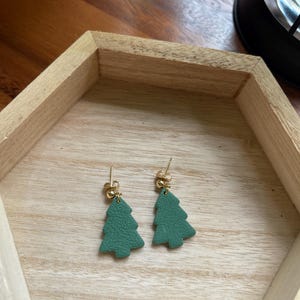 May include: A pair of green Christmas tree earrings with gold-colored posts and accents. The earrings are displayed on a light wooden surface inside a wooden hexagonal tray.